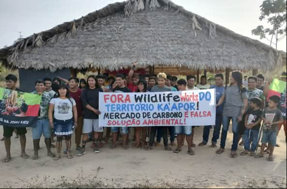 Más de 30 indígenas Ka'apor de pie frente a una casa tradicional con pilares de madera y techo de paja. Las personas sostienen una pancarta de protesta en la que se lee: "¡Fuera Wildlife Works del territorio ka'apor! El mercado de carbono es una falsa sol
