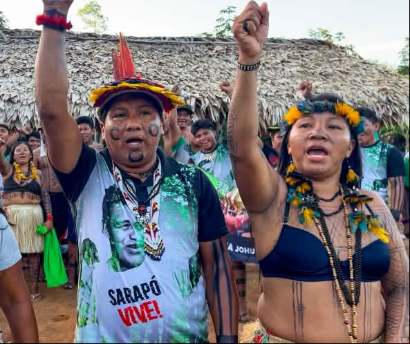 Tres indígenas Ka'apor —dos mujeres y un hombre, con los puños en alto delante de un espacio cubierto con hojas de palma.