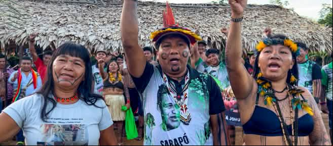 Tres indígenas Ka'apor —dos mujeres y un hombre, con los puños en alto delante de un espacio cubierto con hojas de palma.